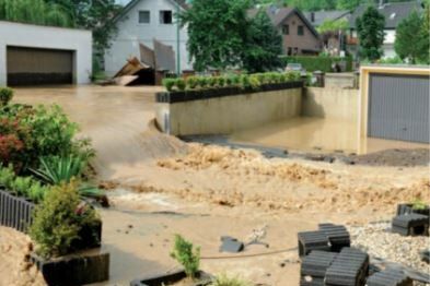 Wohngebiet mit starkem Hochwasser: braune Fluten strömen durch einen Hof und über eine Zufahrt, Garagen und Gartenbereiche sind überflutet